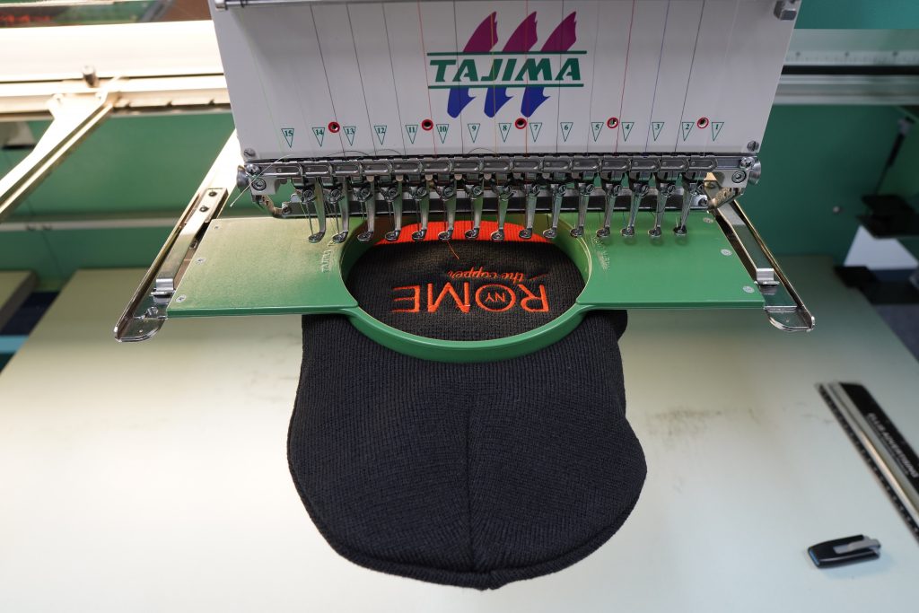 Black and orange beanie on an embroidery machine getting the words "Rome, NY, The Copper City" embroidered on it
