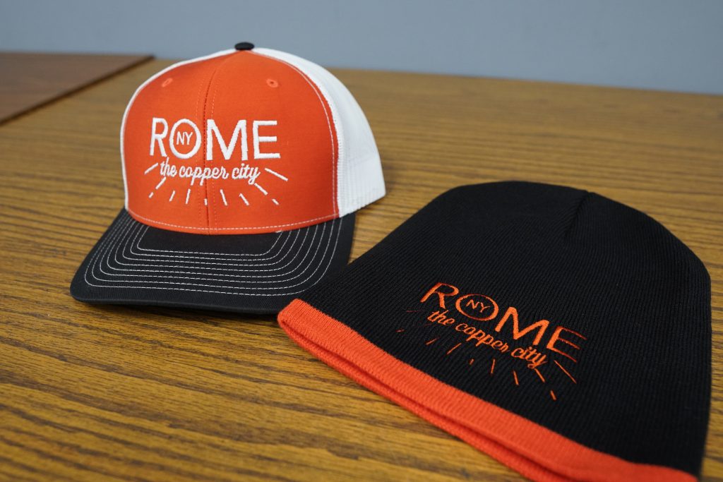 Black and orange beanie and baseball hat with the words "Rome, NY, The Copper City" embroidered into it