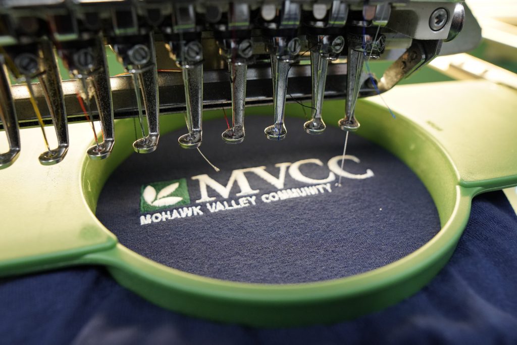 an up close image of an embroidery machine stitching the words MVCC Mohawk Valley Community College into a navy blue t-shirt