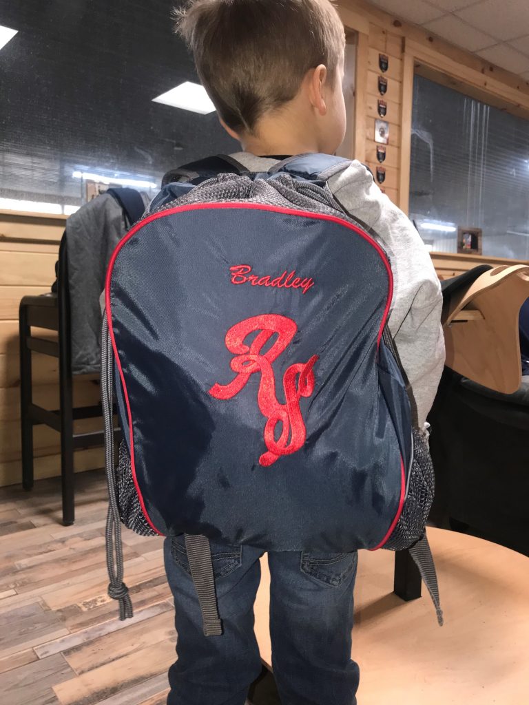 A photo of a little boy with a bookbag on that say his name in red letters, "Bradley" and have the initials RS in red embroidery