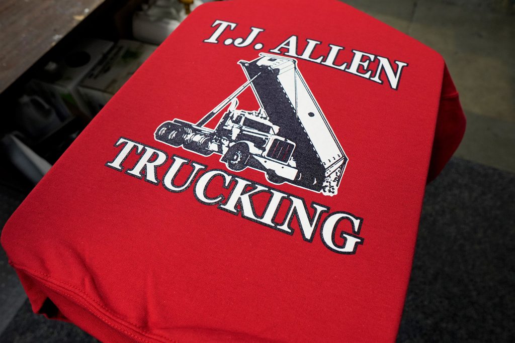 a photo of a red t-shirt getting screen printed. It says T.J. Allen Trucking with a large photo of a truck on the front